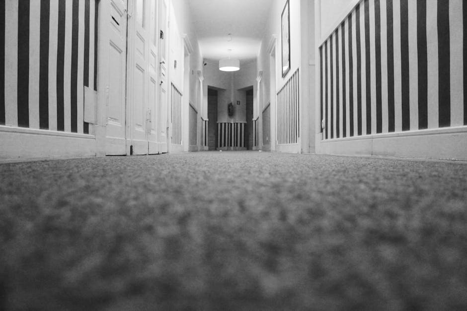 A black-and-white photograph of a clean, well-maintained corridor with a low-angle view from the floor. The corridor has a textured carpet floor that appears freshly vacuumed or deep cleaned, with tex