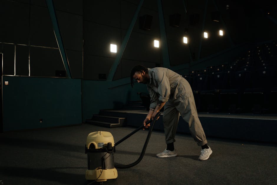 A professional cleaner dressed in a white protective suit, face mask, goggles, and orange gloves is operating a vacuum cleaner or steam cleaning equipment inside a modern living room. The room has lar